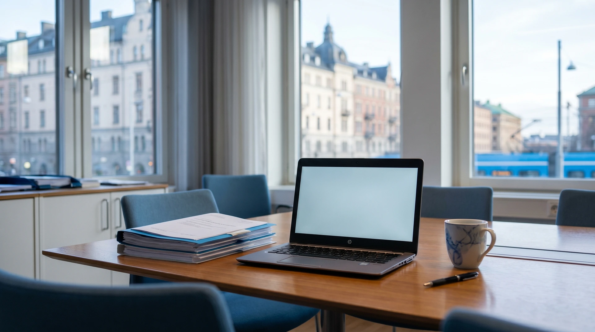 Laptop och pappershögar på kontorsbord med stadsutsikt genom fönster.